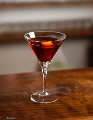 Ruby red Erogin cocktail presented in a martini glass on a wooden bar with a orange twist garnish