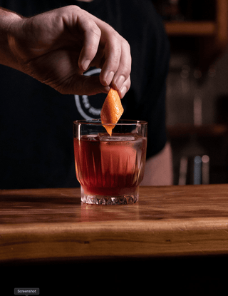 Red Negroni cocktail in a short glass on a wooden bar, a hand places the orange twist garnish 