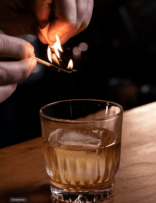 A hand holds a match lightly charring the orange peel garnish over top of a short glass with a large ice cube containing the Ole Fashioned New Zealand Whiskey cocktail