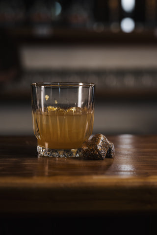 A short glass on a wooden bar holds the penicillin cocktail, dusky orange garnsihed with flakes of ginger. Two whiskey chocolates sit alongside as the perfect mouthful.