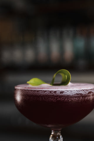 Close up shot of a martini glass with a deep purple Motupipi Martini cocktail, a delicate lime leaf garnishes