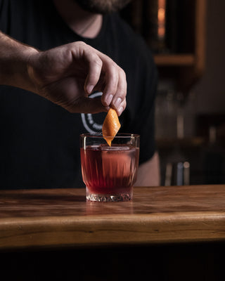Red Negroni cocktail in a short glass on a wooden bar, a hand places the orange twist garnish