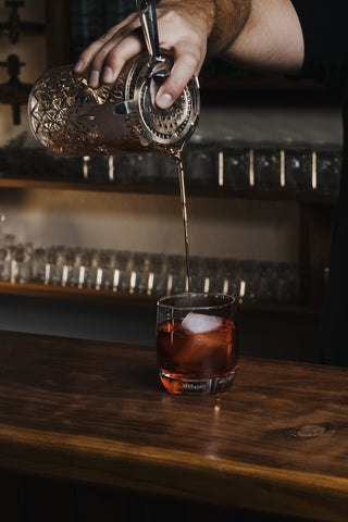 Boulevardier cocktail on wooden bar, large square ice cube with ruby red drink being poured from a cocktail jug