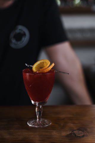 A twist of orange oil is the last step to this deep red cocktail made with Boysenberry Liqueur and Delightful gin, garnished with orange on a metal cocktail spoon