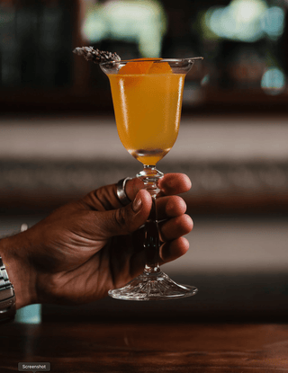 Hand holds a vibrant yellow cocktail in a wine glass garnished with a lavender flower