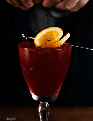 A twist of orange oil is the last step to this deep red cocktail made with Boysenberry Liqueur and Delightful gin, garnished with orange on a metal cocktail spoon