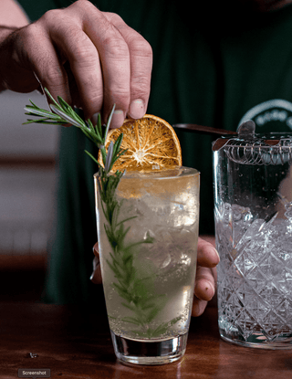 Tall glass with a over sized rosemary sprig filled with ice and Naranjo cocktail, a hand places the finishing touch of a dried orange round into the drink