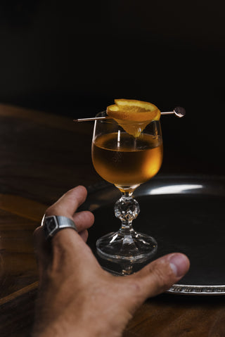 A hand reaches for the Orange Blossom cocktail, a stunning glass holds orange liquid garnished with fresh orange round