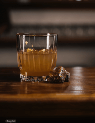 A short glass on a wooden bar holds the penicillin cocktail, dusky orange garnsihed with flakes of ginger. Two whiskey chocolates sit alongside as the perfect mouthful. 