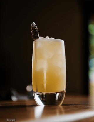 A tall glass holds the pohara sunrise, a yellow cocktail garnsihed with lavender