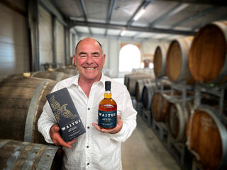 Terry smiling with a bottle of Waitui Whiskey in front of the aging Whiskey barrels