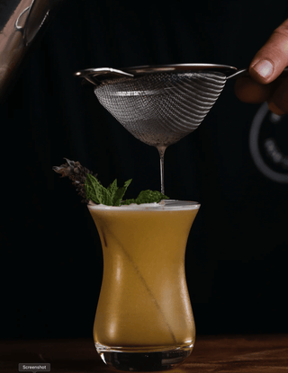 The last drops of the Botanist are starined into the glass, a dusky yellow cocktail garnished with mint and lavender