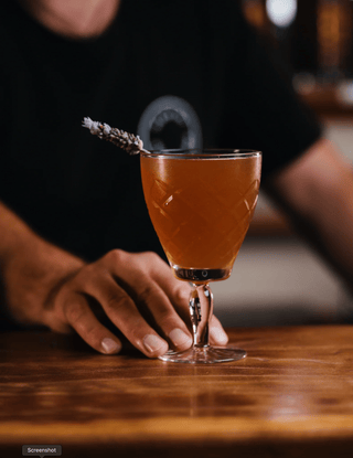 Jeremy passes a dusky orange cocktail garnished with lavender across the bar 
