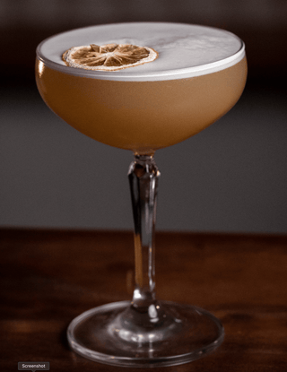 A martini glass holds the Twisted Whiskey Sour, a delicate layer of foam tops the yellow cocktail, garnished with dried lemon
