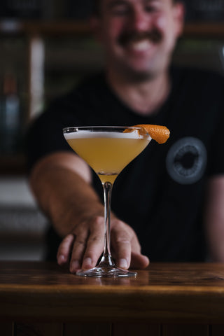 Jeremy passes a glass towards you of Delightful Tangelo Sour, vibrant orange cocktail in a martini glass with an orange twist garnish