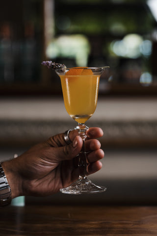 Hand holds a vibrant yellow cocktail in a wine glass garnished with a lavender flower