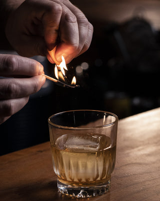 A hand holds a match lightly charring the orange peel garnish over top of a short glass with a large ice cube containing the Ole Fashioned New Zealand Whiskey cocktail