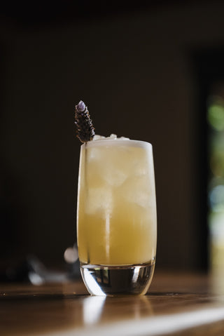 A tall glass holds the pohara sunrise, a yellow cocktail garnsihed with lavender