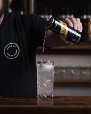 Soda water is poured from a height into a tall glass of Tom Collins on a wooden bar