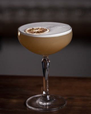 A martini glass holds the Twisted Whiskey Sour, a delicate layer of foam tops the yellow cocktail, garnished with dried lemon