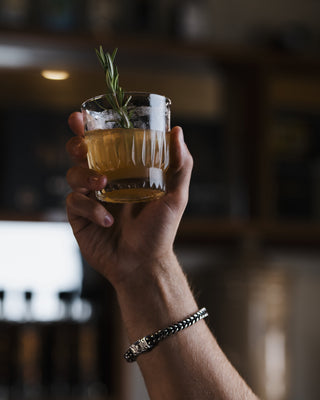 A hold holds the Vegan Lamb Shank in the air, a short glass of orange spirit garnished with a rosemary sprig